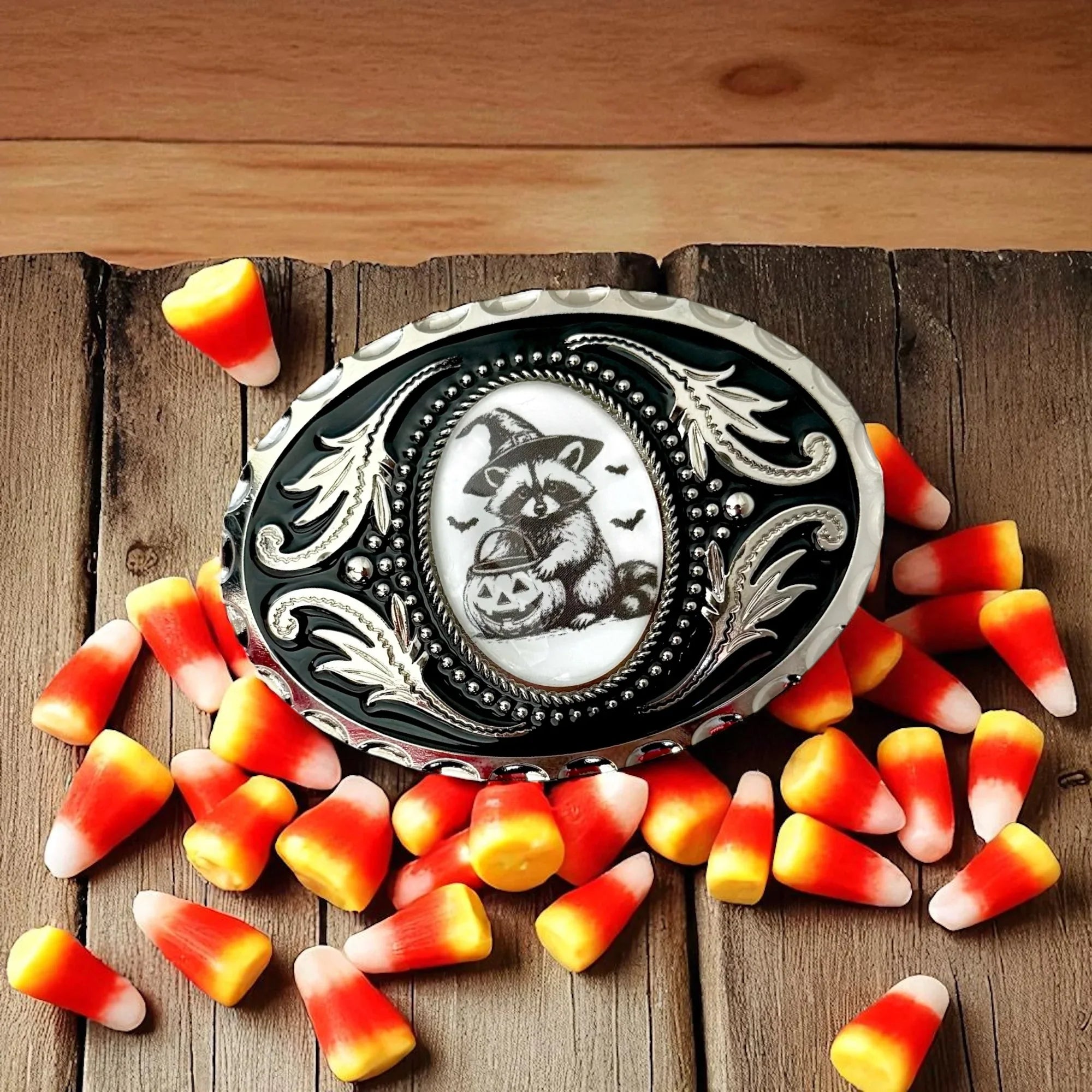 Raccoon Belt buckle with witch outfit surrounded by candy corn on a wooden surface