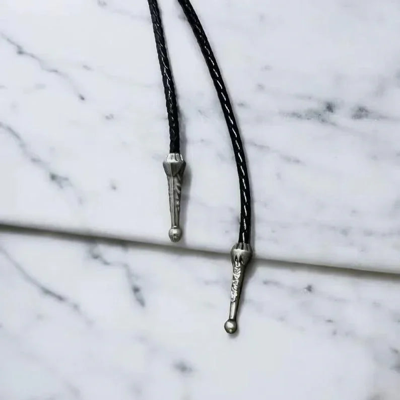 Infographic from The Bold Bow Tie titled Bolo Tie Features. Right: A vintage-style bolo tie with a blue and white cowboy riding a horse illustration inside an ornate bronze filigree frame on a black braided cord. Left: Lines connect the image to three checkmark features: Handmade, 39" Long Cord, and Pendant is 1.5" (L) x 1.25" (W). Bottom text reads Perfect Addition to Your Outfit.