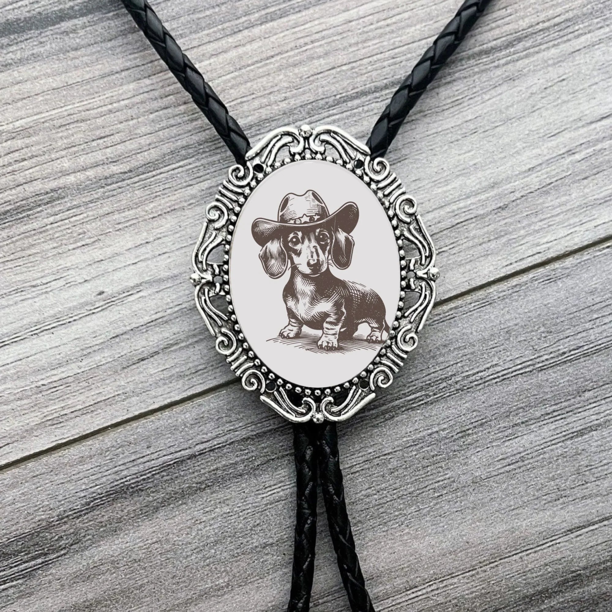 Infographic from The Bold Bow Tie titled Bolo Tie Features. Right: A vintage-style bolo tie with a blue and white cowboy riding a horse illustration inside an ornate bronze filigree frame on a black braided cord. Left: Lines connect the image to three checkmark features: Handmade, 39" Long Cord, and Pendant is 1.5" (L) x 1.25" (W). Bottom text reads Perfect Addition to Your Outfit.