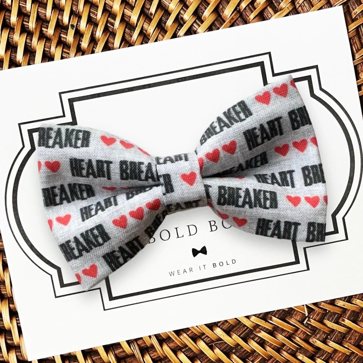 Valentine’s Day dog Bow tie with 'Heart Breaker' text and red hearts on a white card with brand name, placed on a woven surface.