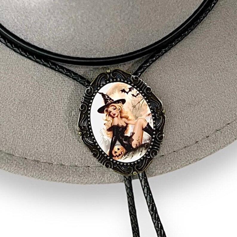 Oval pendant showing a blonde witch lounging in a black outfit beside a carved pumpkin with bats in a spooky backdrop.