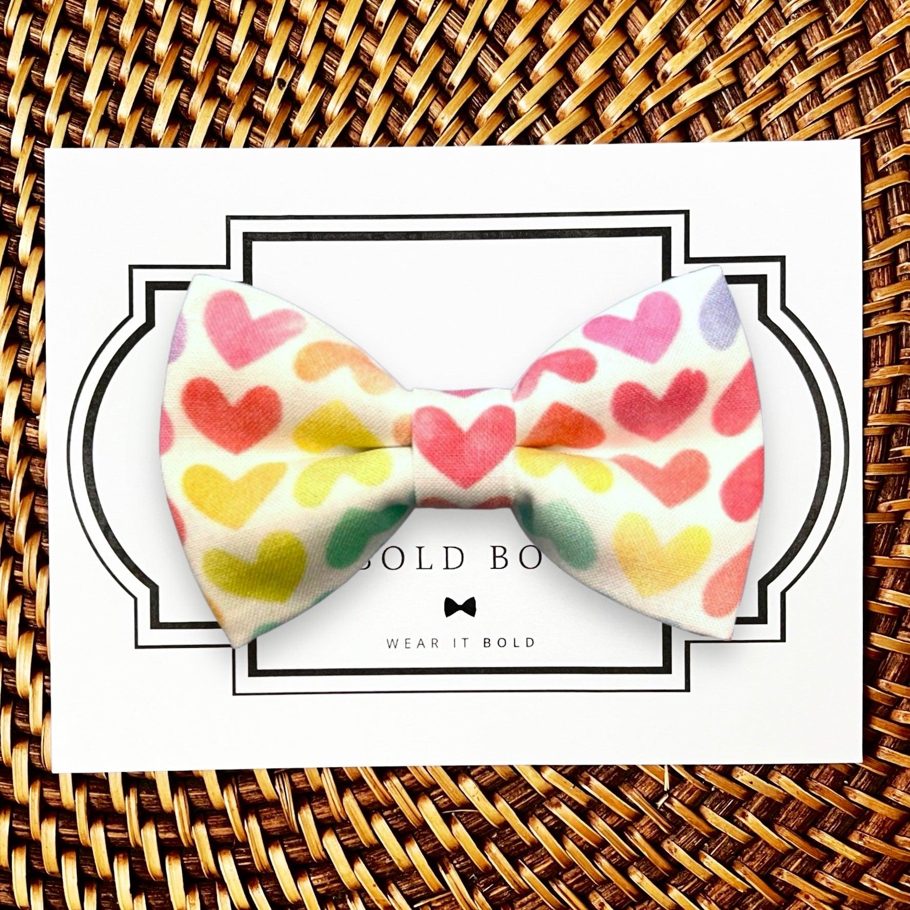 Colorful heart dog bow tie on a white card with 'The Bold Bow Tie’ branding, placed on a woven surface.