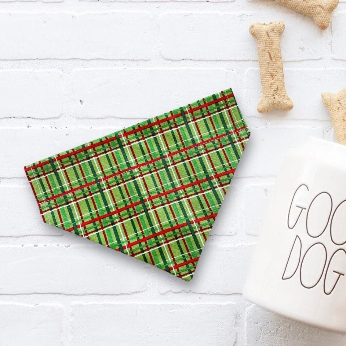 Green plaid Christmas dog bandana on a white surface with 'Good Dog' mug and dog bones.