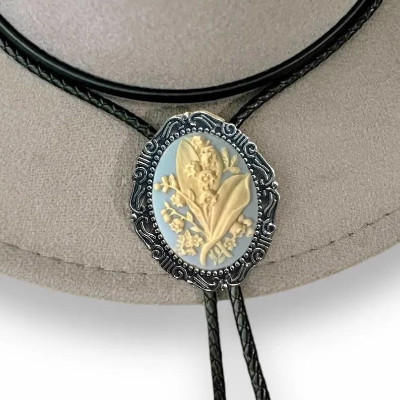 Infographic from The Bold Bow Tie titled Bolo Tie Features. Right: A vintage-style bolo tie with a blue and white cowboy riding a horse illustration inside an ornate bronze filigree frame on a black braided cord. Left: Lines connect the image to three checkmark features: Handmade, 39" Long Cord, and Pendant is 1.5" (L) x 1.25" (W). Bottom text reads Perfect Addition to Your Outfit.