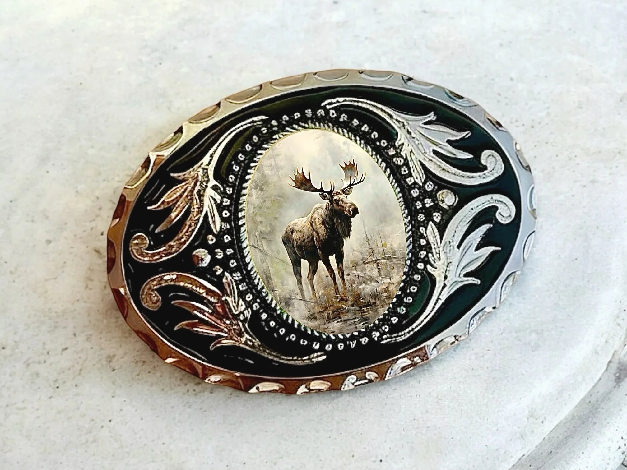Decorative cowboy belt buckle with a moose motif surrounded by ornate black and gold scrollwork.