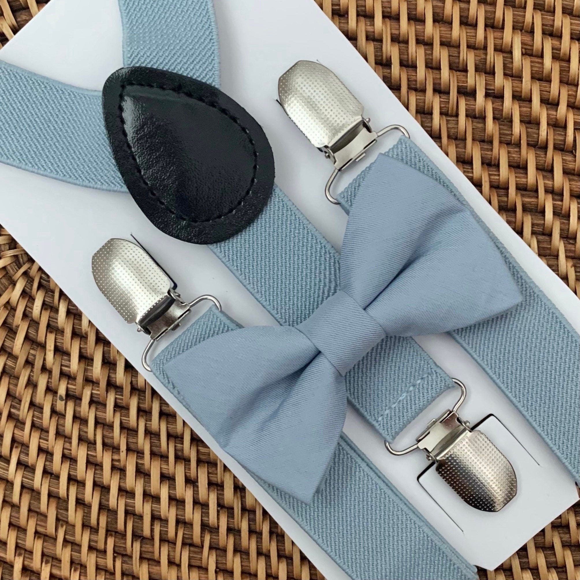 Dusty blue bow tie with silver clips on dusty blue suspenders for groomsmen, ring bearer outfit and wedding on a woven surface
