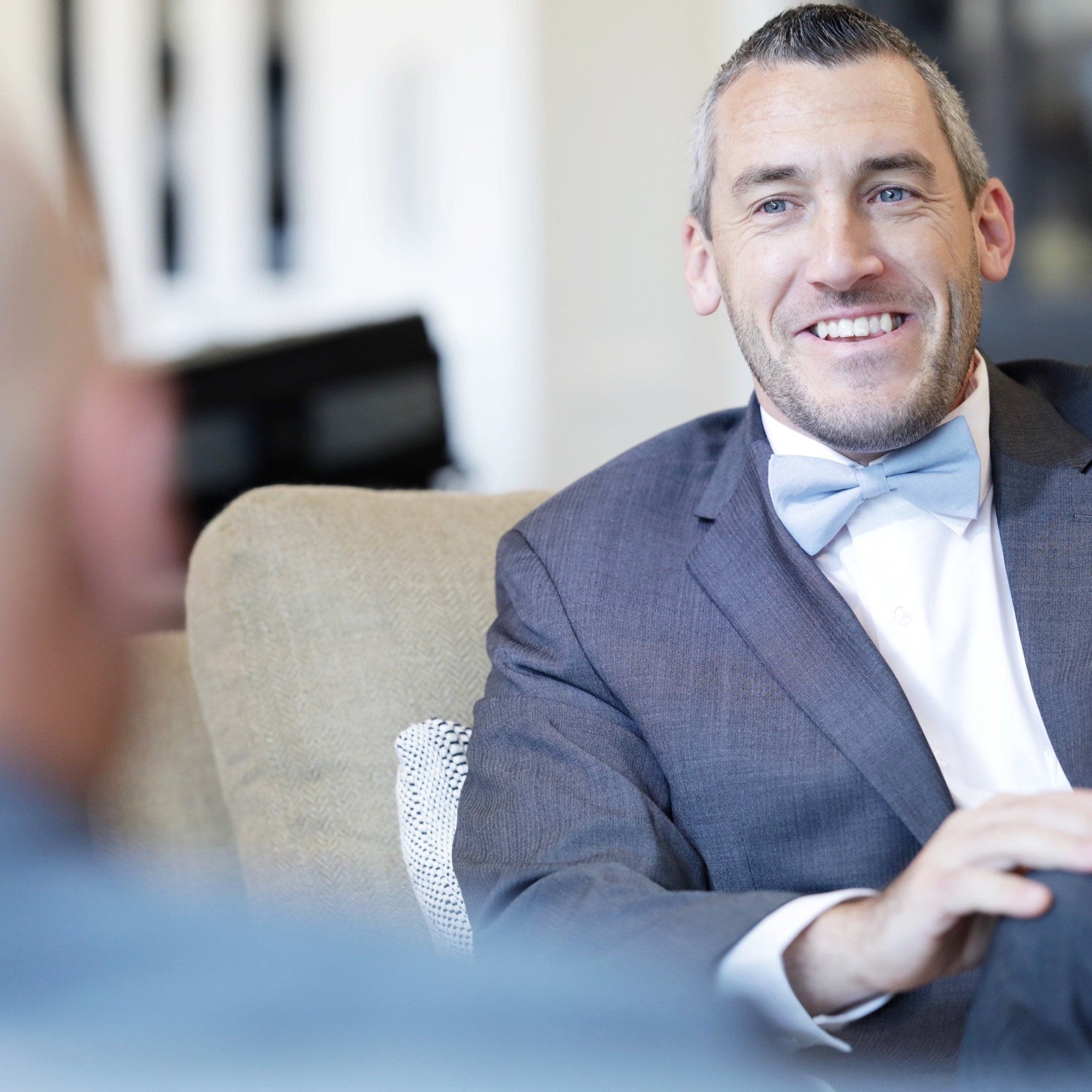 Man in a suit and dusty blue bow tie sitting on a couch, smiling.