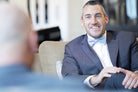 Man in a suit and dusty blue bow tie sitting on a couch, smiling.