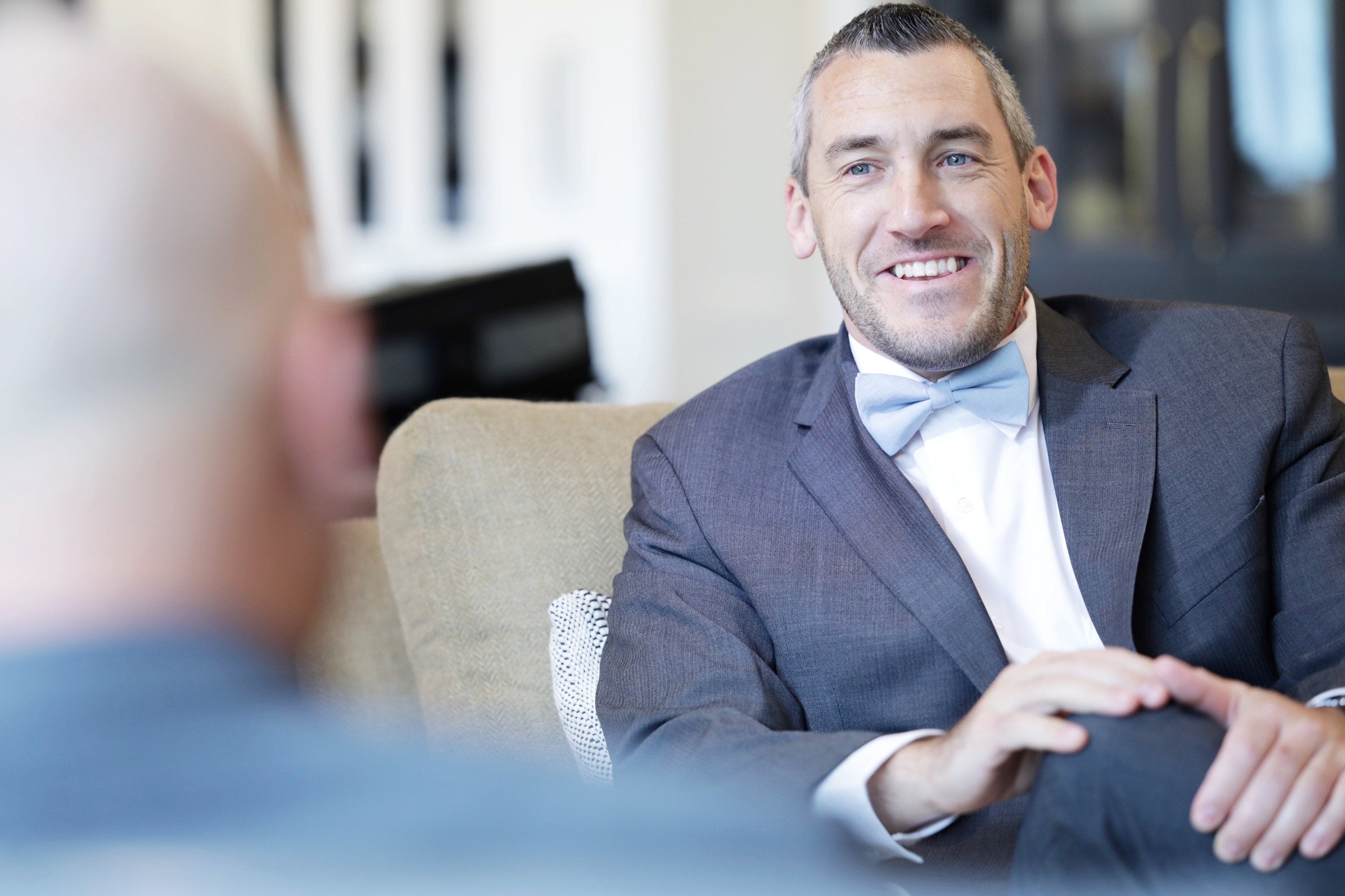 Man in a suit and dusty blue bow tie sitting on a couch, smiling.