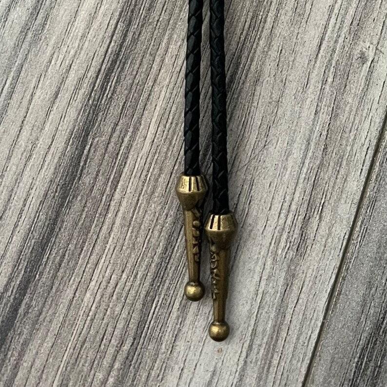 Infographic from The Bold Bow Tie titled Bolo Tie Features. Right: A vintage-style bolo tie with a blue and white cowboy riding a horse illustration inside an ornate bronze filigree frame on a black braided cord. Left: Lines connect the image to three checkmark features: Handmade, 39" Long Cord, and Pendant is 1.5" (L) x 1.25" (W). Bottom text reads Perfect Addition to Your Outfit.