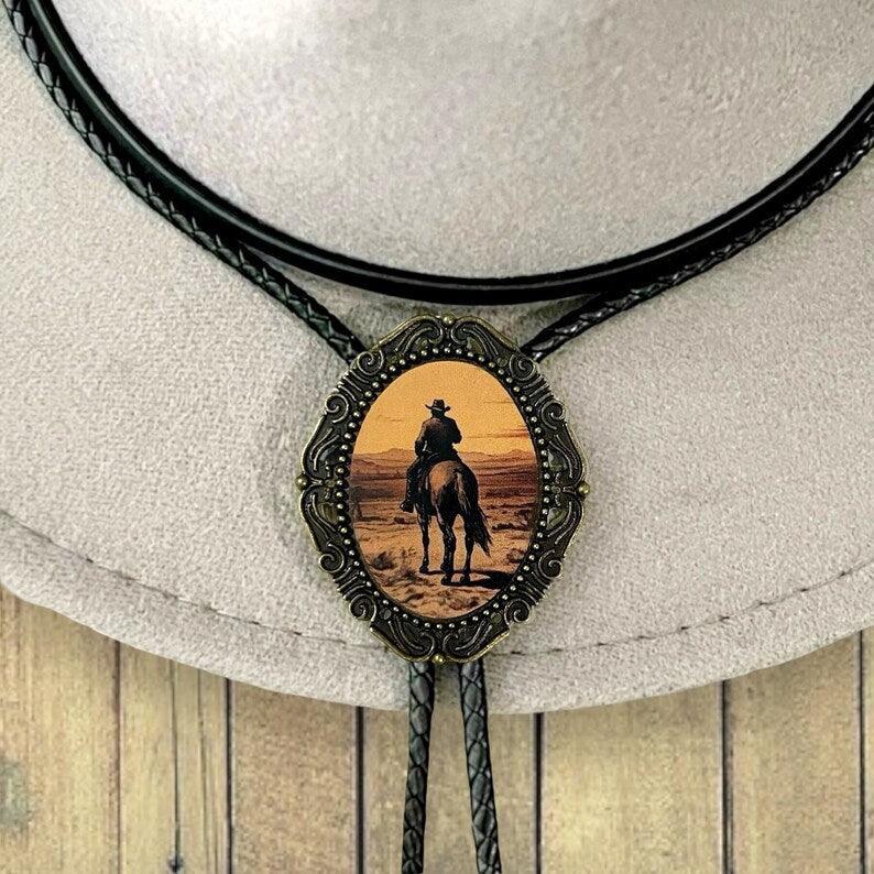 Infographic from The Bold Bow Tie titled Bolo Tie Features. Right: A vintage-style bolo tie with a blue and white cowboy riding a horse illustration inside an ornate bronze filigree frame on a black braided cord. Left: Lines connect the image to three checkmark features: Handmade, 39" Long Cord, and Pendant is 1.5" (L) x 1.25" (W). Bottom text reads Perfect Addition to Your Outfit.