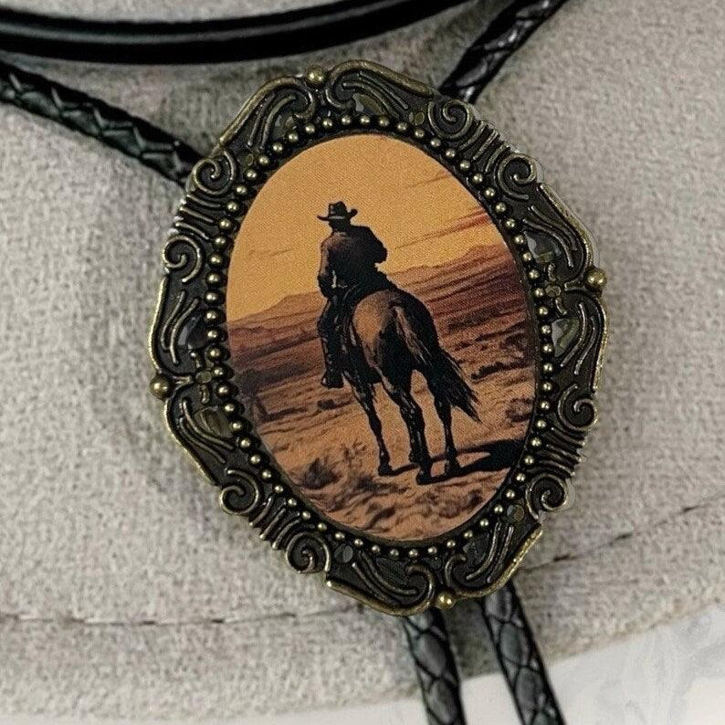 Sunset Cowboy Bolo Tie with ornate bronze frame surrounding an oval desert scene of a cowboy on horseback.