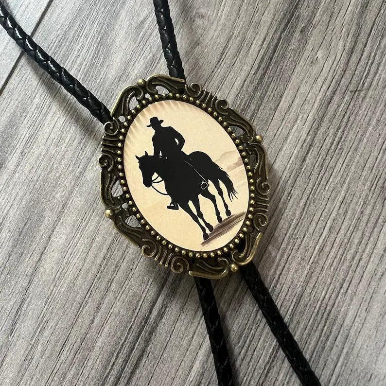 Infographic from The Bold Bow Tie titled Bolo Tie Features. Right: A vintage-style bolo tie with a blue and white cowboy riding a horse illustration inside an ornate bronze filigree frame on a black braided cord. Left: Lines connect the image to three checkmark features: Handmade, 39" Long Cord, and Pendant is 1.5" (L) x 1.25" (W). Bottom text reads Perfect Addition to Your Outfit.