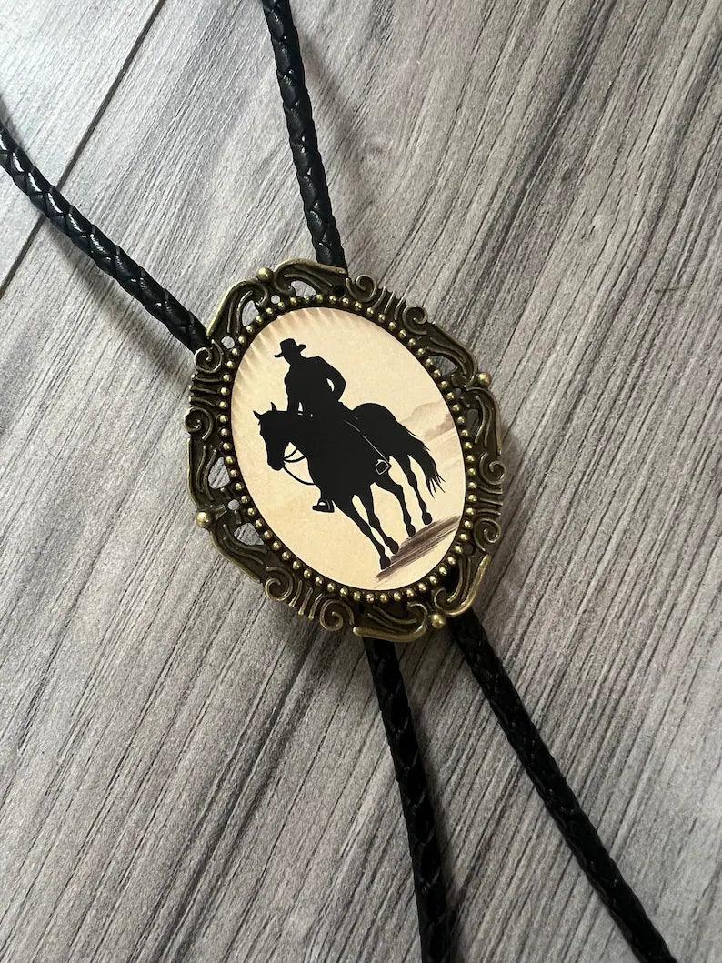 Infographic from The Bold Bow Tie titled Bolo Tie Features. Right: A vintage-style bolo tie with a blue and white cowboy riding a horse illustration inside an ornate bronze filigree frame on a black braided cord. Left: Lines connect the image to three checkmark features: Handmade, 39" Long Cord, and Pendant is 1.5" (L) x 1.25" (W). Bottom text reads Perfect Addition to Your Outfit.