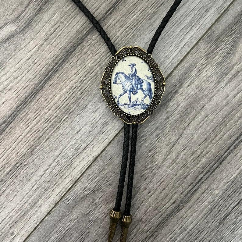 Infographic from The Bold Bow Tie titled Bolo Tie Features. Right: A vintage-style bolo tie with a blue and white cowboy riding a horse illustration inside an ornate bronze filigree frame on a black braided cord. Left: Lines connect the image to three checkmark features: Handmade, 39" Long Cord, and Pendant is 1.5" (L) x 1.25" (W). Bottom text reads Perfect Addition to Your Outfit.