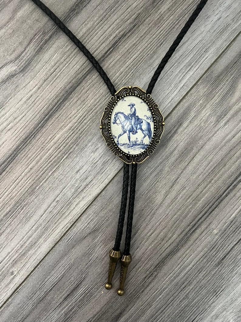 Infographic from The Bold Bow Tie titled Bolo Tie Features. Right: A vintage-style bolo tie with a blue and white cowboy riding a horse illustration inside an ornate bronze filigree frame on a black braided cord. Left: Lines connect the image to three checkmark features: Handmade, 39" Long Cord, and Pendant is 1.5" (L) x 1.25" (W). Bottom text reads Perfect Addition to Your Outfit.