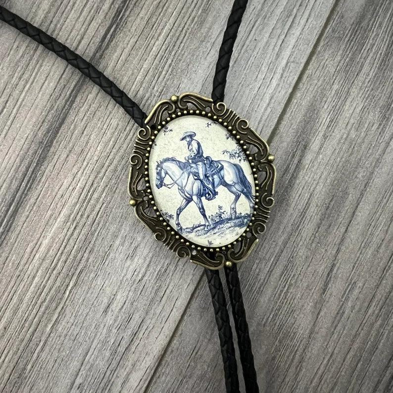 Cowboy bolo tie with an ornate bronze frame surrounding a blue and white porcelain rider centerpiece on black braided cord.