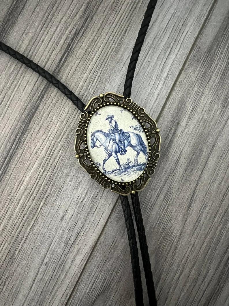 Cowboy bolo tie with an ornate bronze frame surrounding a blue and white porcelain rider centerpiece on black braided cord.