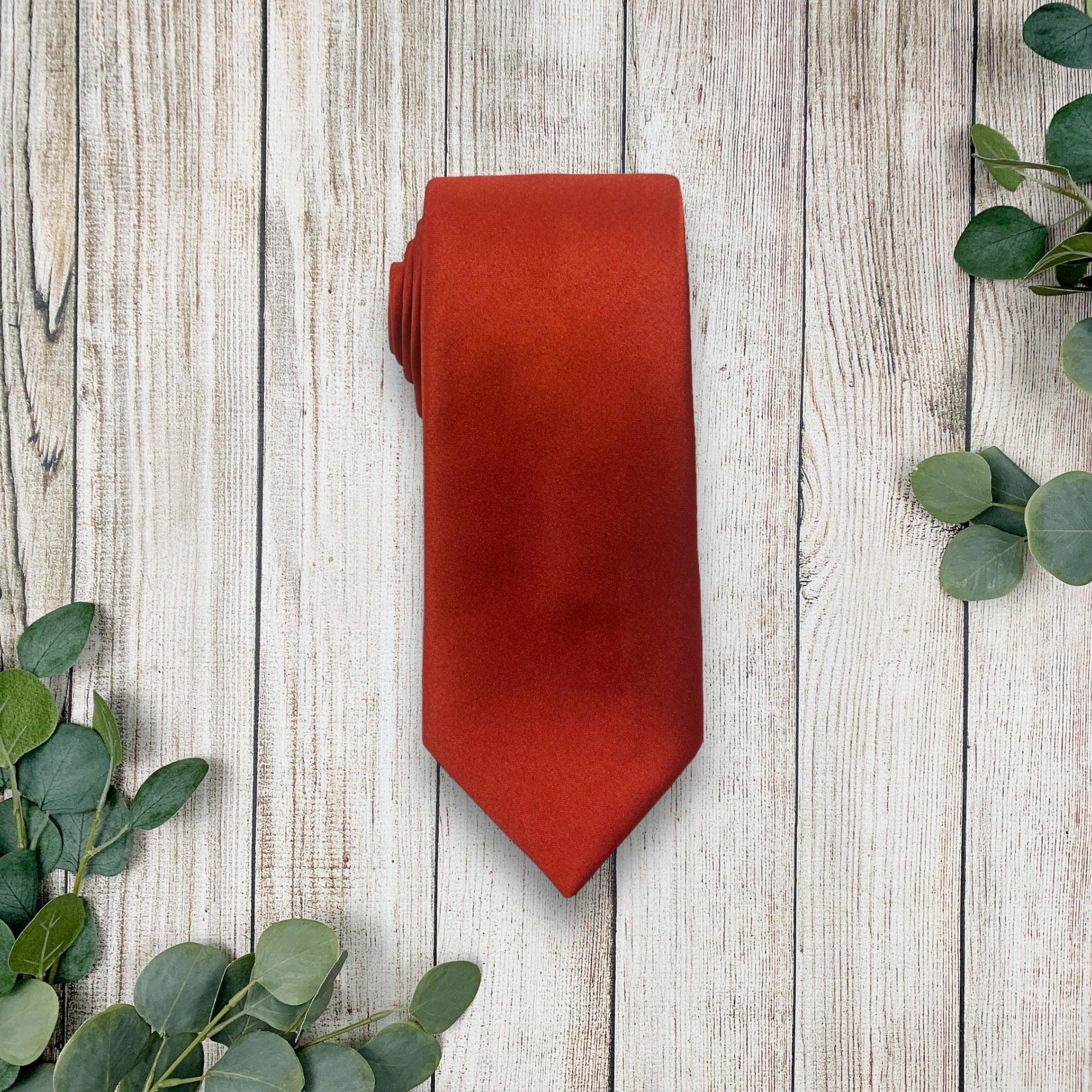 Burnt orange satin necktie folded on a rustic wood surface with eucalyptus leaves