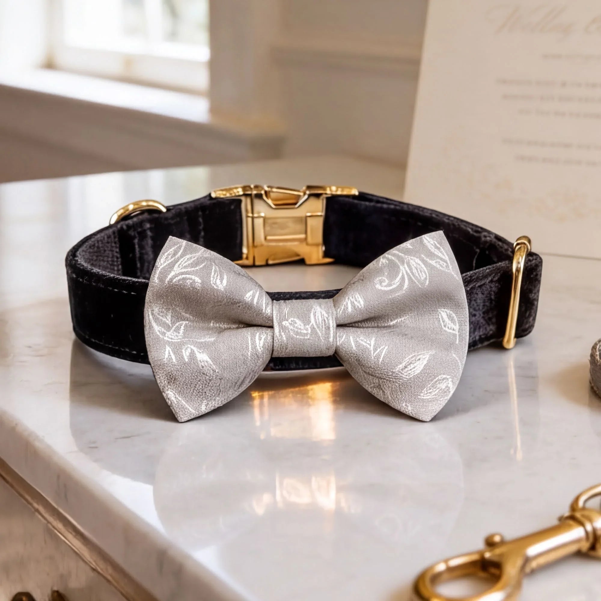 A black velvet dog collar with a gold buckle and a white bow tie with a floral pattern are displayed on a marble countertop.