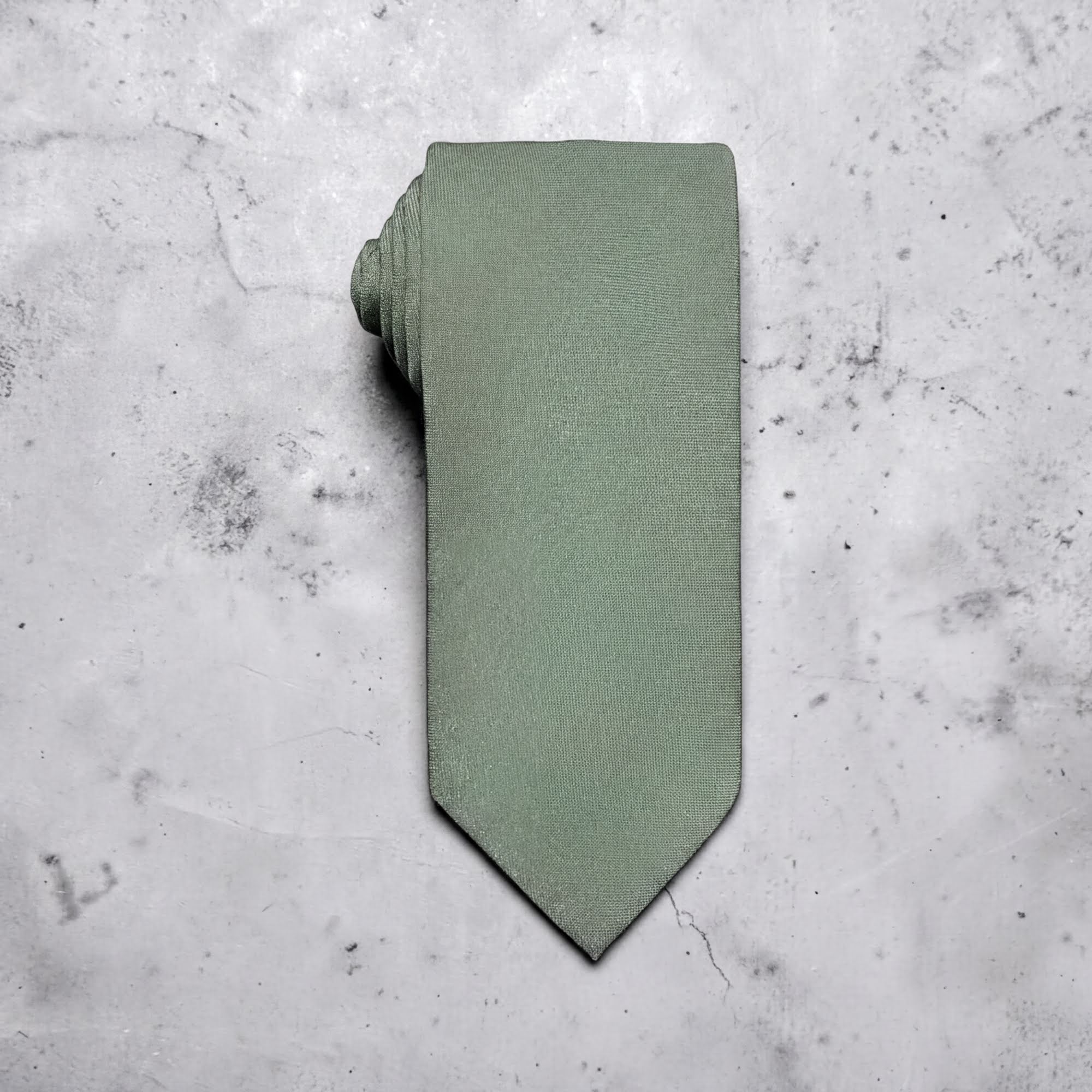 A sage green tie for men’s wedding necktie with a matching fabric neck loop, laid flat on a textured light gray concrete surface.
