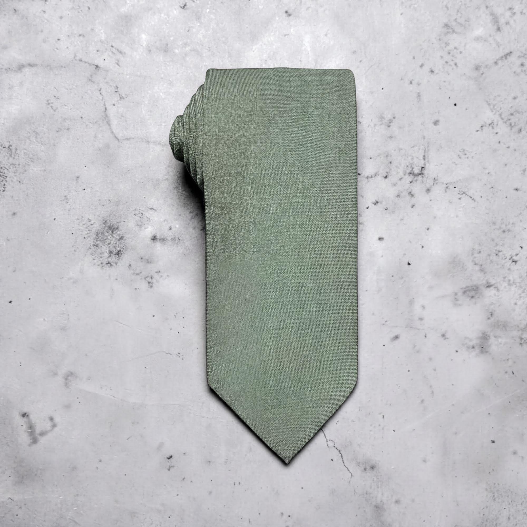 A sage green tie for men’s wedding necktie with a matching fabric neck loop, laid flat on a textured light gray concrete surface.