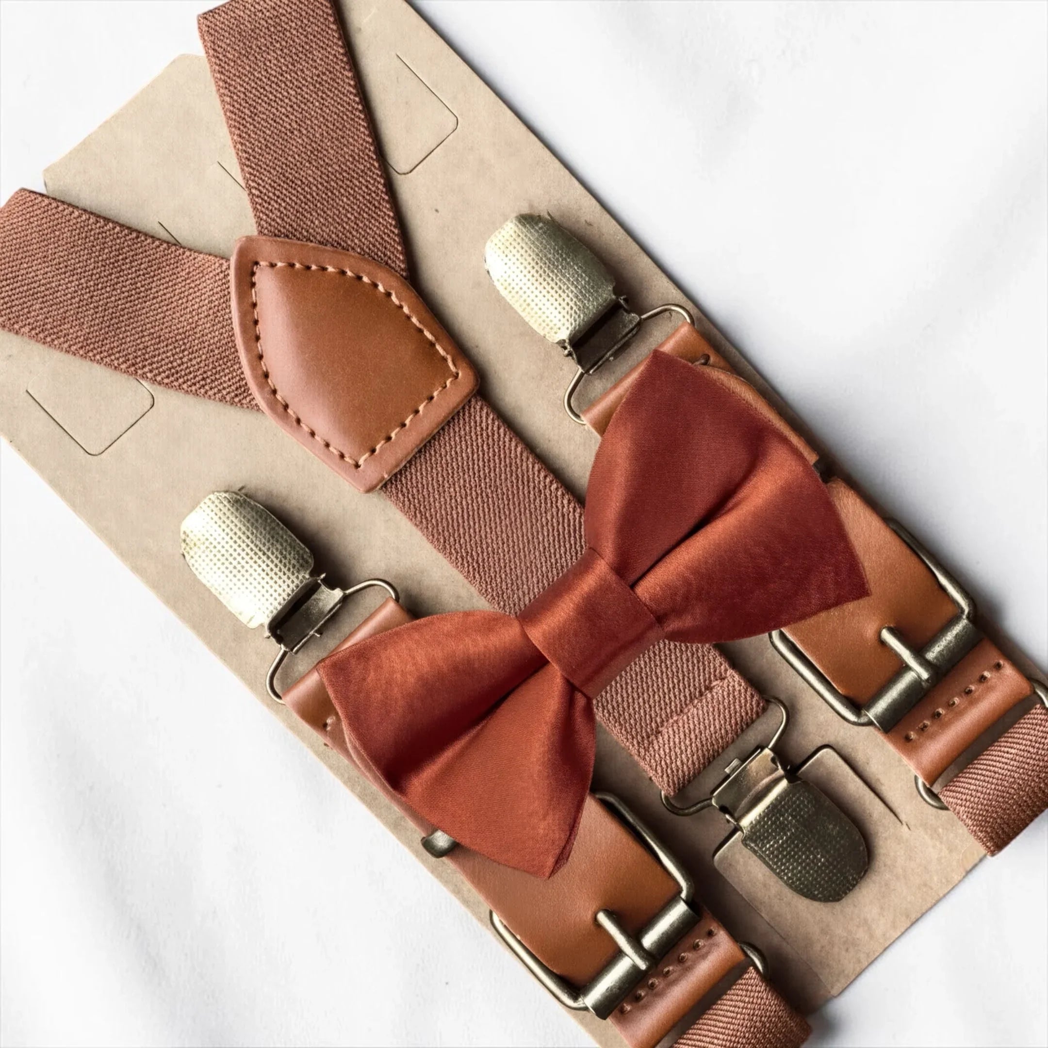 A package containing brown suspenders with a red bow tie attached, placed on a white surface.