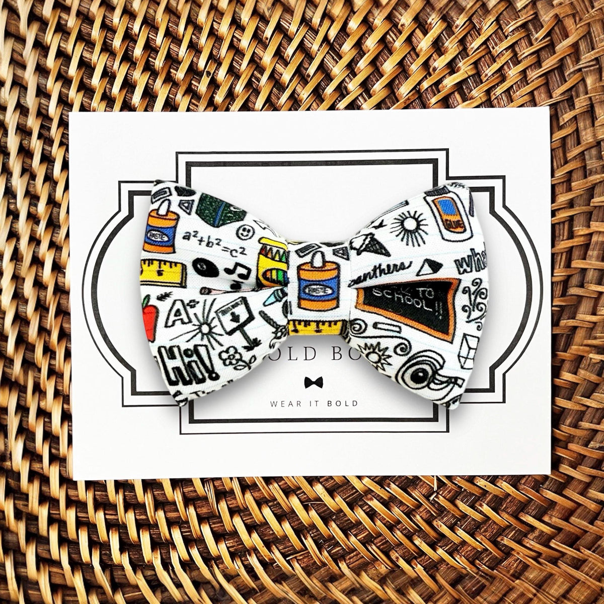 A bow tie with a pattern of school-related items, such as a chalkboard, calculator, and pencils, printed on it. The bow tie is placed on a white card with a black border.
