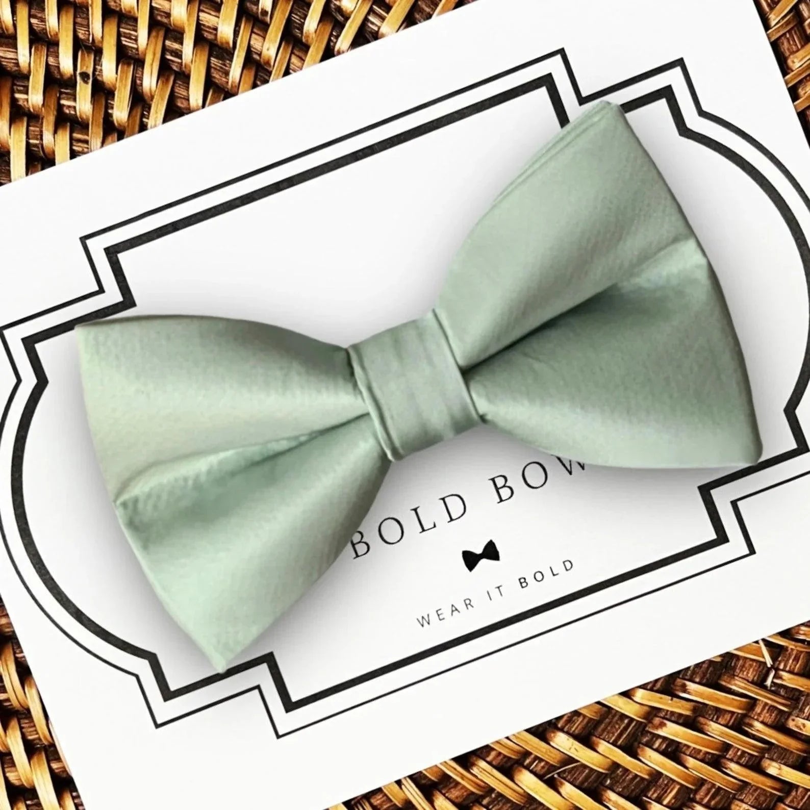 Sage green satin bow tie on a white display card with a black decorative border on a woven surface