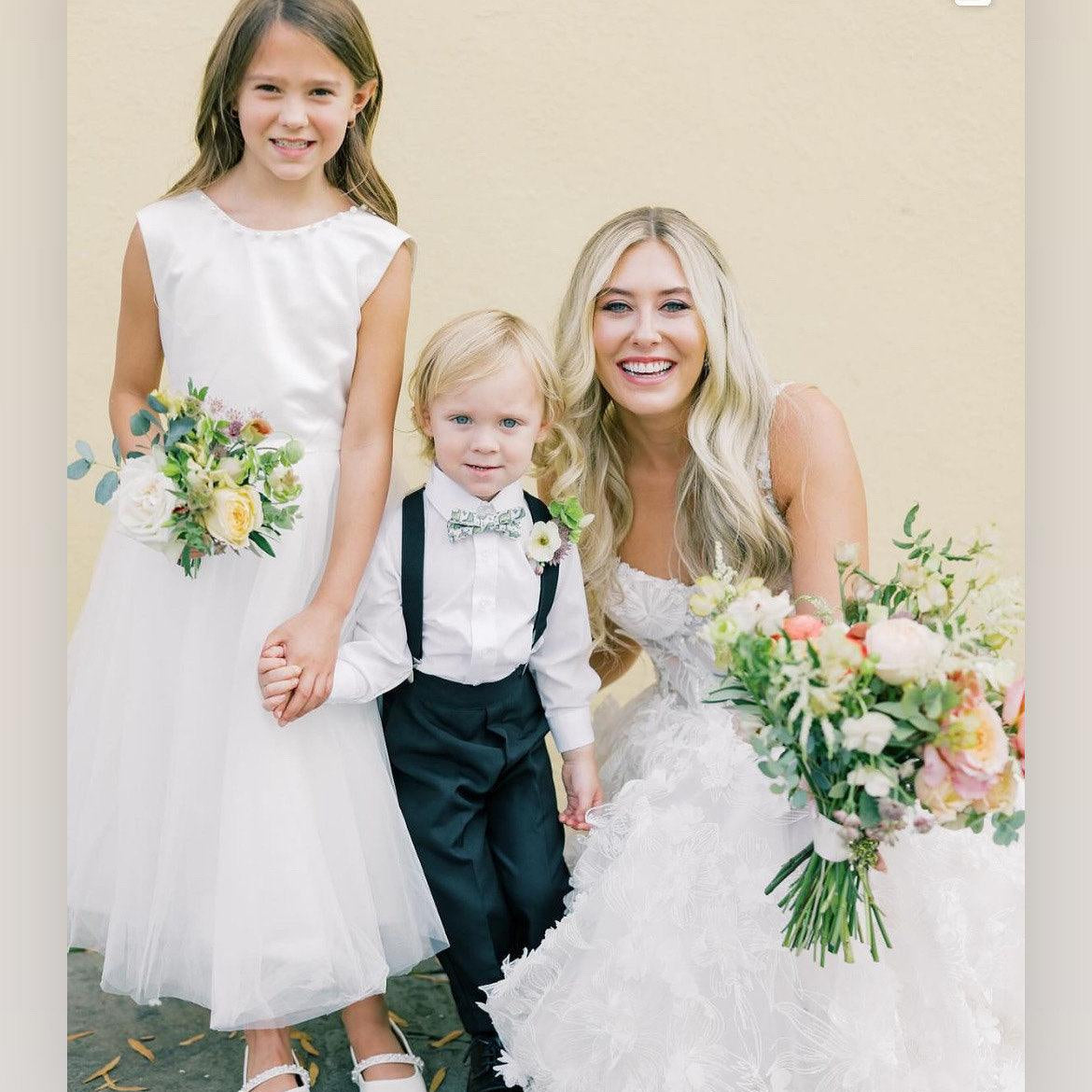 Ring bearer wearing sage green dinosaur bow tie at a wedding surrounded by the bride and flower girl.