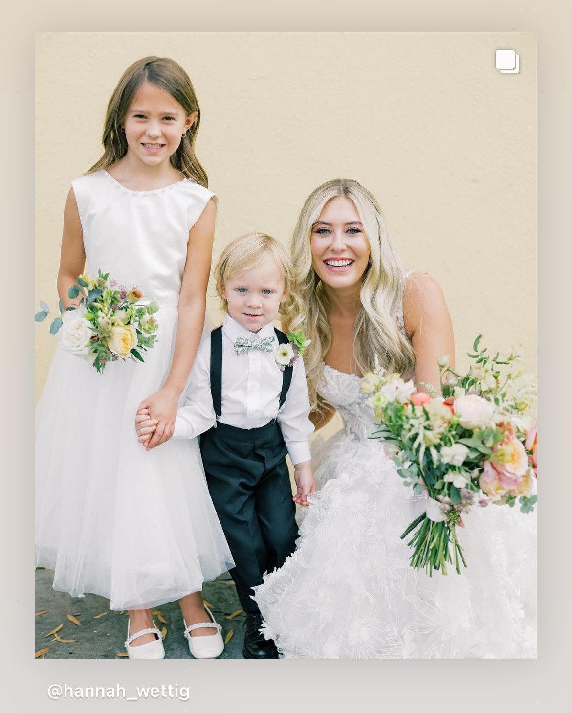 Ring bearer wearing sage green dinosaur bow tie at a wedding surrounded by the bride and flower girl.