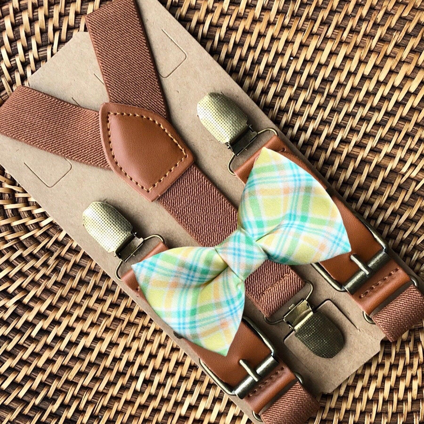 Pastel plaid bow tie with cognac suspenders displayed on a leather strap over a brown textured backdrop