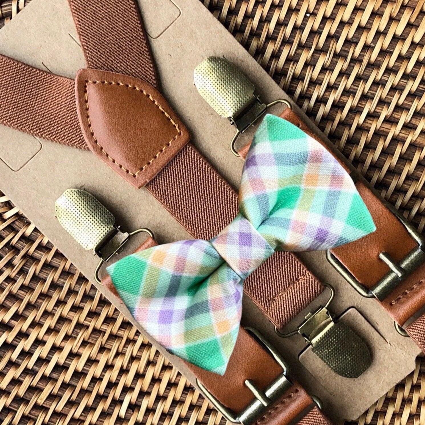 Green and purple plaid Easter bow tie paired with cognac suspenders on a tan leather strap against a woven backdrop.
