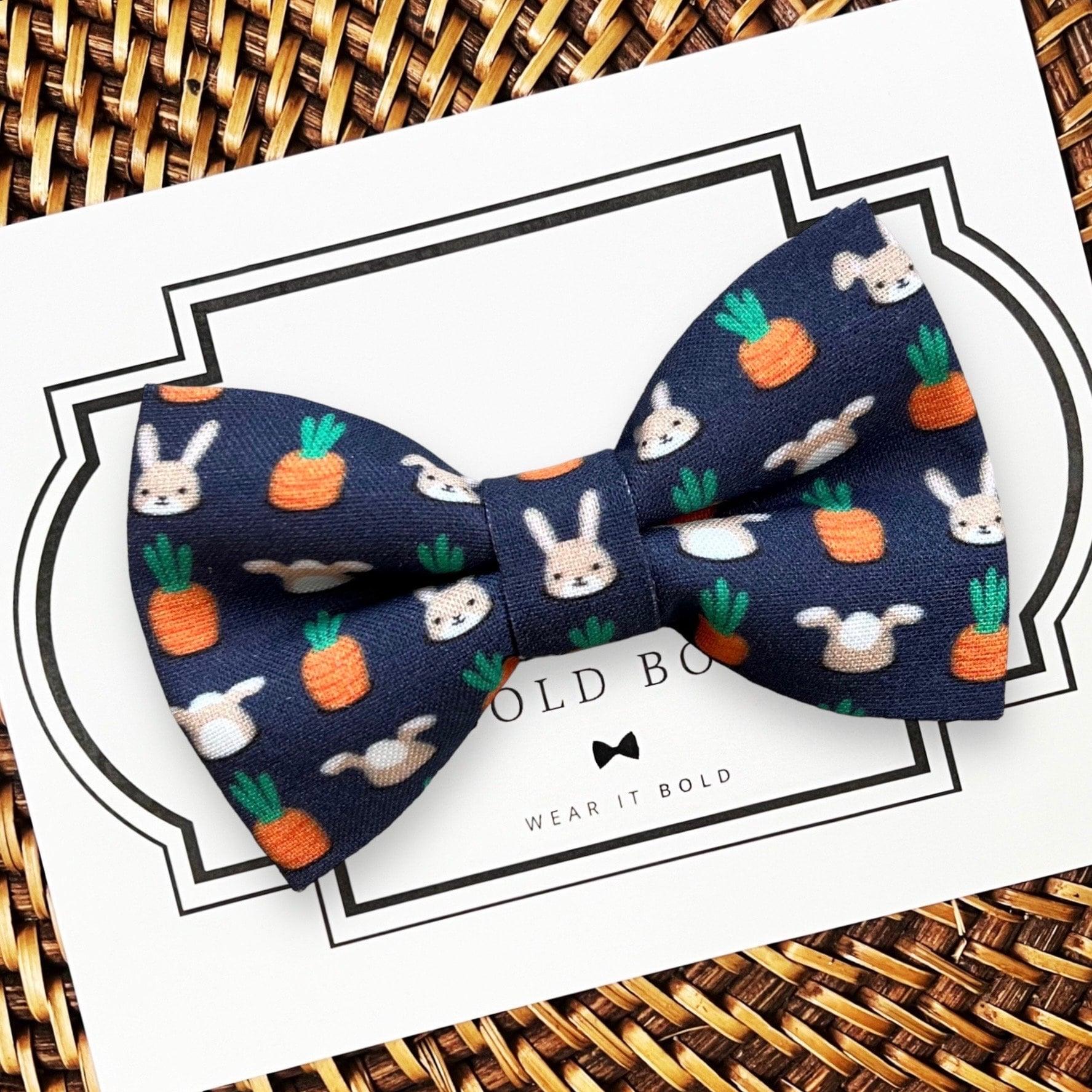 A navy blue dog bow tie with a pattern of white rabbits and orange carrots, displayed on a card with the text 'The Bold Bow Tie, Wear it Bold'.