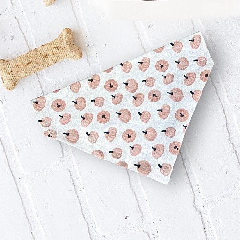 Flat lay of a pink pumpkin patterned dog bandana on a white textured surface with a bone toy nearby