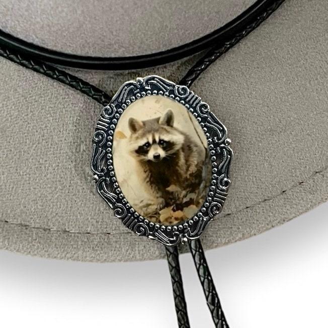 Infographic from The Bold Bow Tie titled Bolo Tie Features. Right: A vintage-style bolo tie with a blue and white cowboy riding a horse illustration inside an ornate bronze filigree frame on a black braided cord. Left: Lines connect the image to three checkmark features: Handmade, 39" Long Cord, and Pendant is 1.5" (L) x 1.25" (W). Bottom text reads Perfect Addition to Your Outfit.