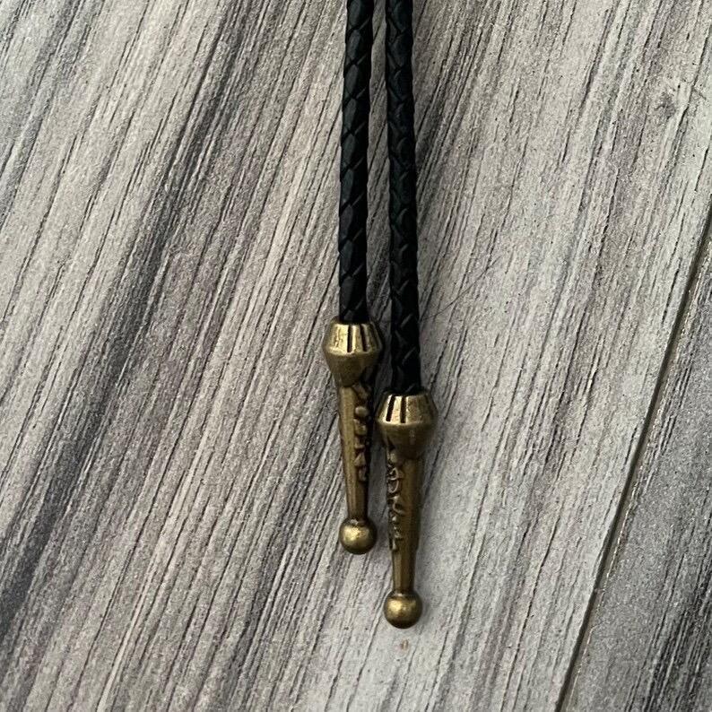 Bolo tie size chart from The Bold Bow Tie. The image displays a braided leather bolo tie with a round silver concho-style slide and silver metal tips, set against a multi-colored striped fabric background. A vertical dotted arrow on the right spans the full length of the piece, accompanied by text indicating a 39 inch cord length.