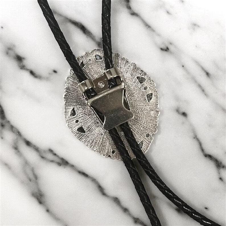 Close-up of a silver circular concho bolo tie with a decorative buckle on black cords against a marble surface.