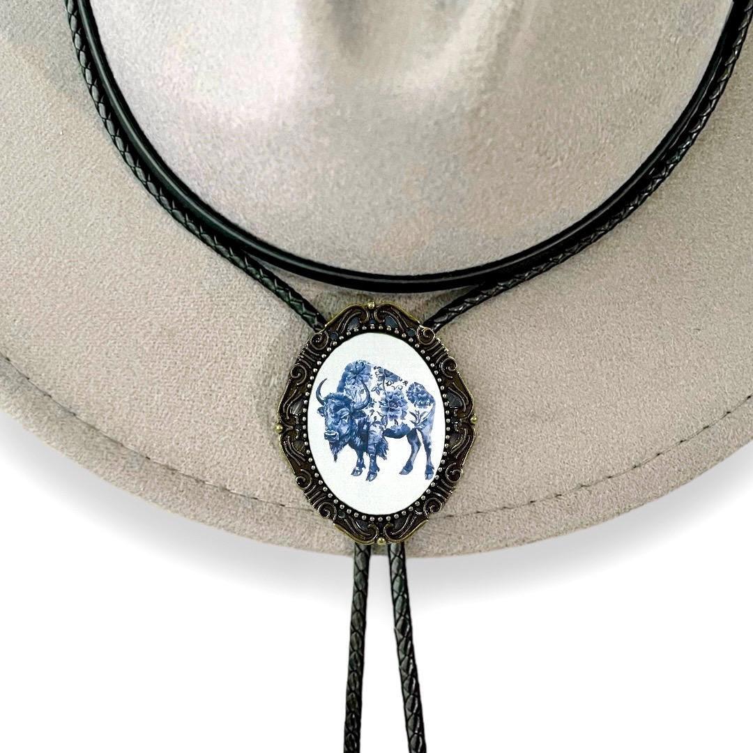 Infographic from The Bold Bow Tie titled Bolo Tie Features. Right: A vintage-style bolo tie with a blue and white cowboy riding a horse illustration inside an ornate bronze filigree frame on a black braided cord. Left: Lines connect the image to three checkmark features: Handmade, 39" Long Cord, and Pendant is 1.5" (L) x 1.25" (W). Bottom text reads Perfect Addition to Your Outfit.