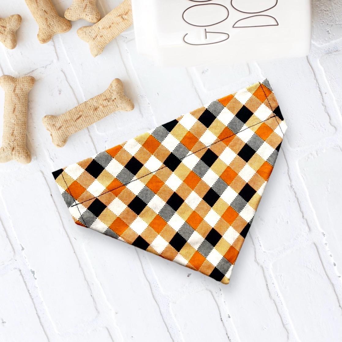 Orange plaid Halloween dog bandana on a white surface with bone shaped dog biscuits nearby