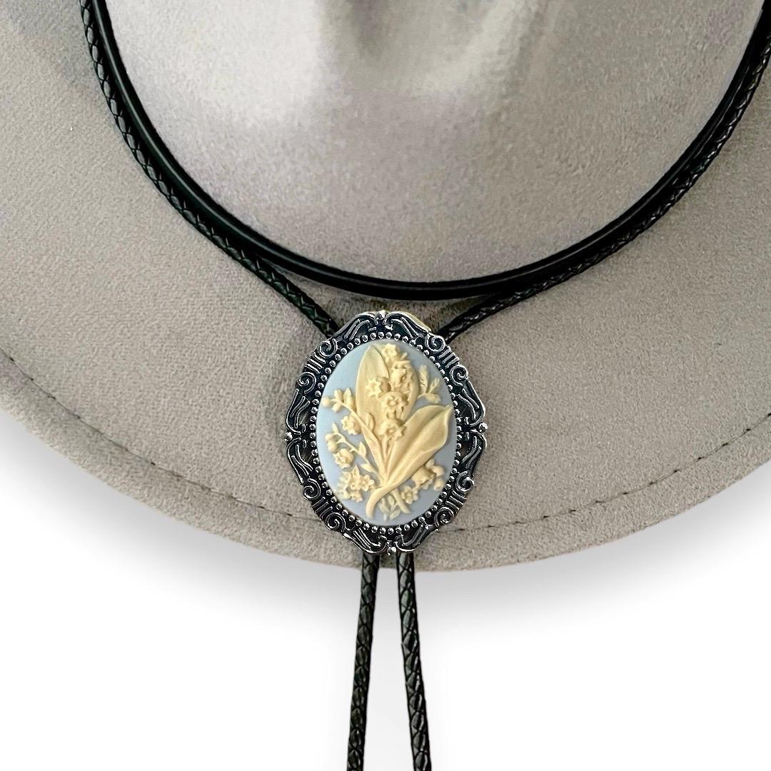 Infographic from The Bold Bow Tie titled Bolo Tie Features. Right: A vintage-style bolo tie with a blue and white cowboy riding a horse illustration inside an ornate bronze filigree frame on a black braided cord. Left: Lines connect the image to three checkmark features: Handmade, 39" Long Cord, and Pendant is 1.5" (L) x 1.25" (W). Bottom text reads Perfect Addition to Your Outfit.