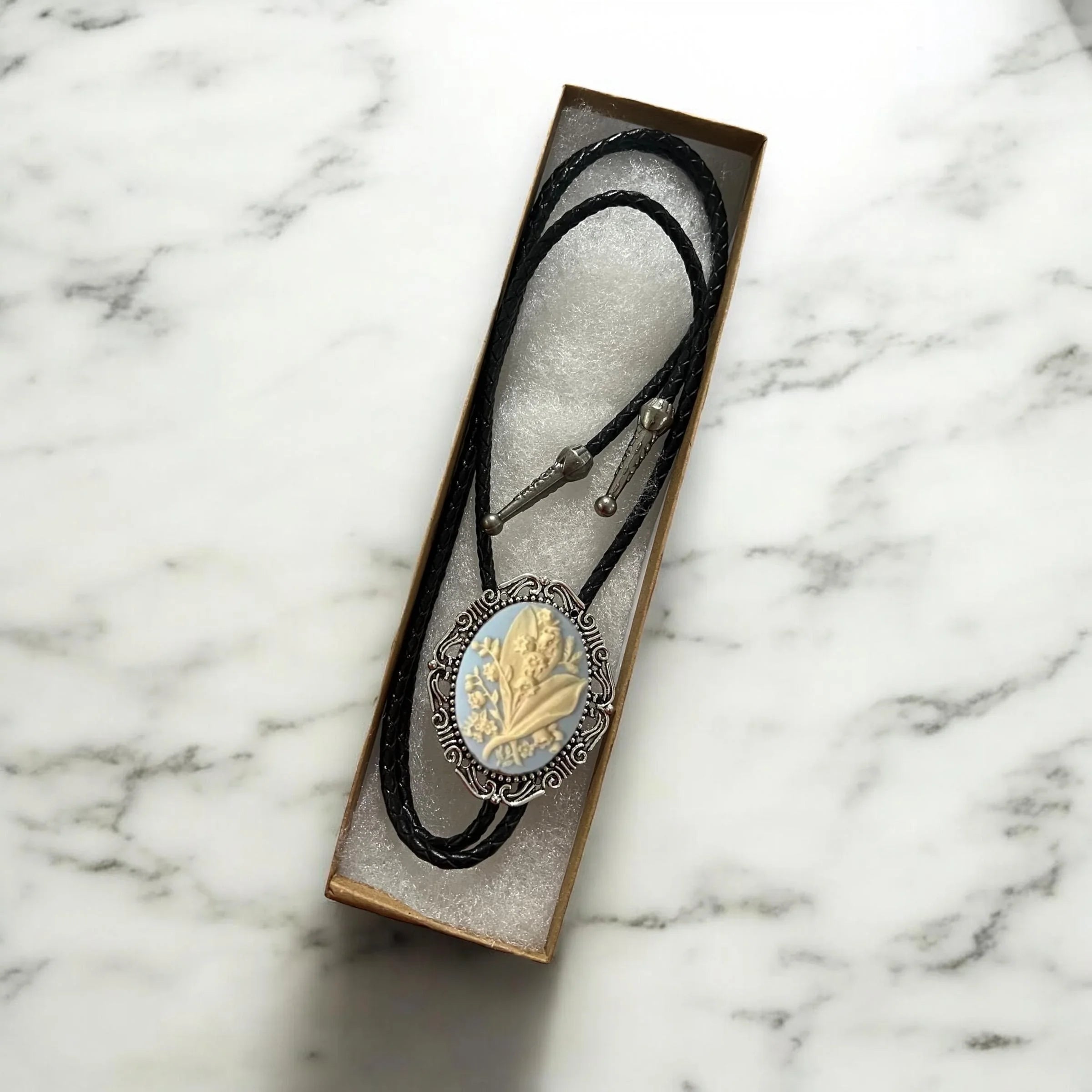 Bolo tie size chart from The Bold Bow Tie. The image displays a braided leather bolo tie with a round silver concho-style slide and silver metal tips, set against a multi-colored striped fabric background. A vertical dotted arrow on the right spans the full length of the piece, accompanied by text indicating a 39 inch cord length.