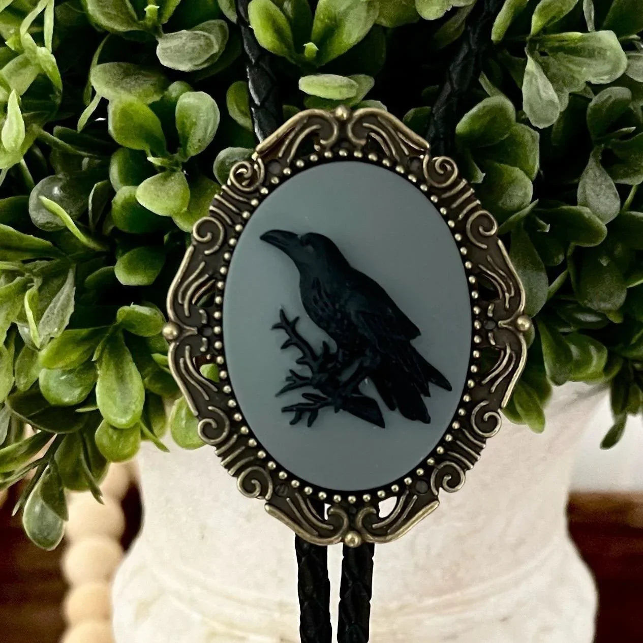 Infographic from The Bold Bow Tie titled Bolo Tie Features. Right: A vintage-style bolo tie with a blue and white cowboy riding a horse illustration inside an ornate bronze filigree frame on a black braided cord. Left: Lines connect the image to three checkmark features: Handmade, 39" Long Cord, and Pendant is 1.5" (L) x 1.25" (W). Bottom text reads Perfect Addition to Your Outfit.