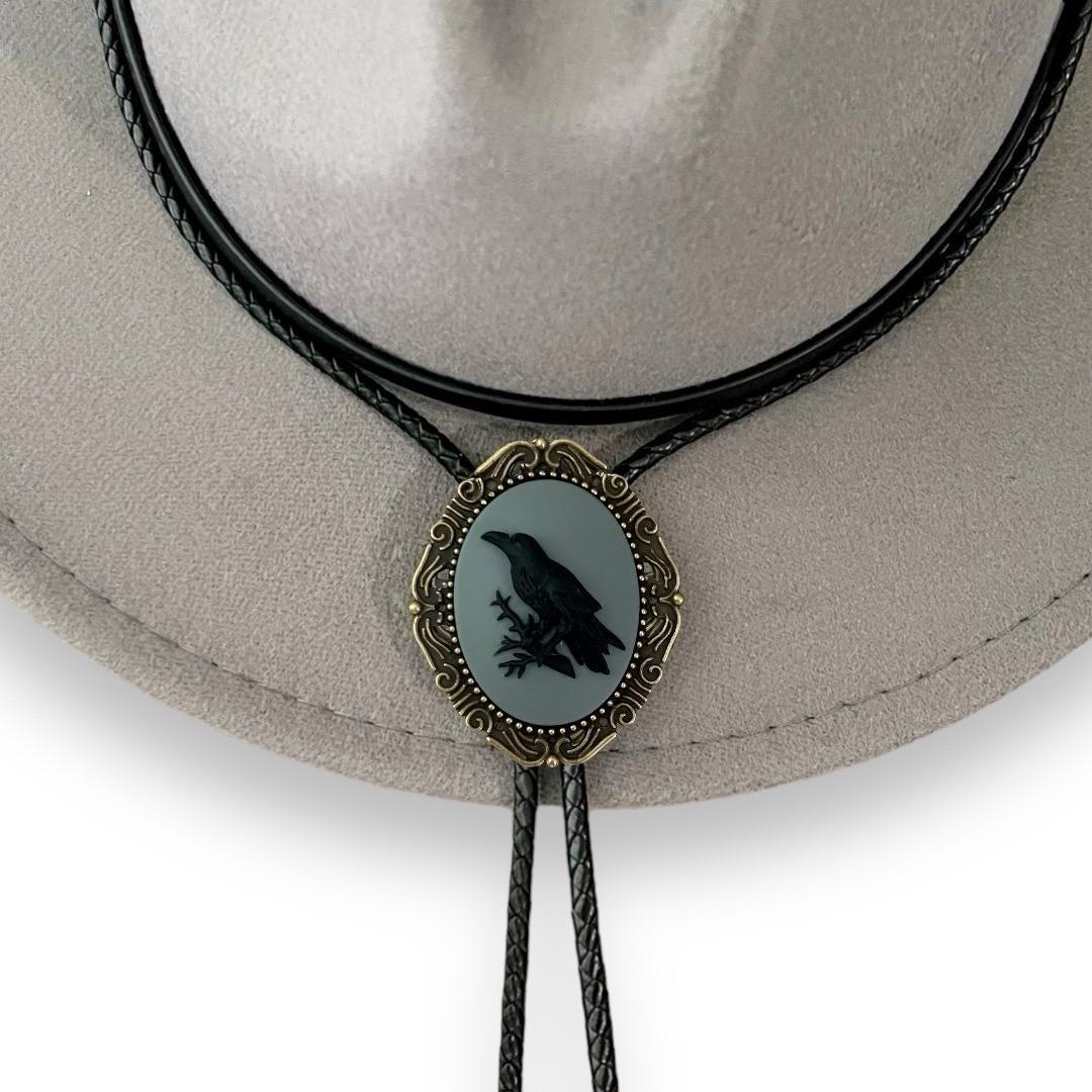 Crow Bolo tie centered on a light hat featuring a gray oval centerpiece with crow silhouette, ornate brass surround and braided leather cords.