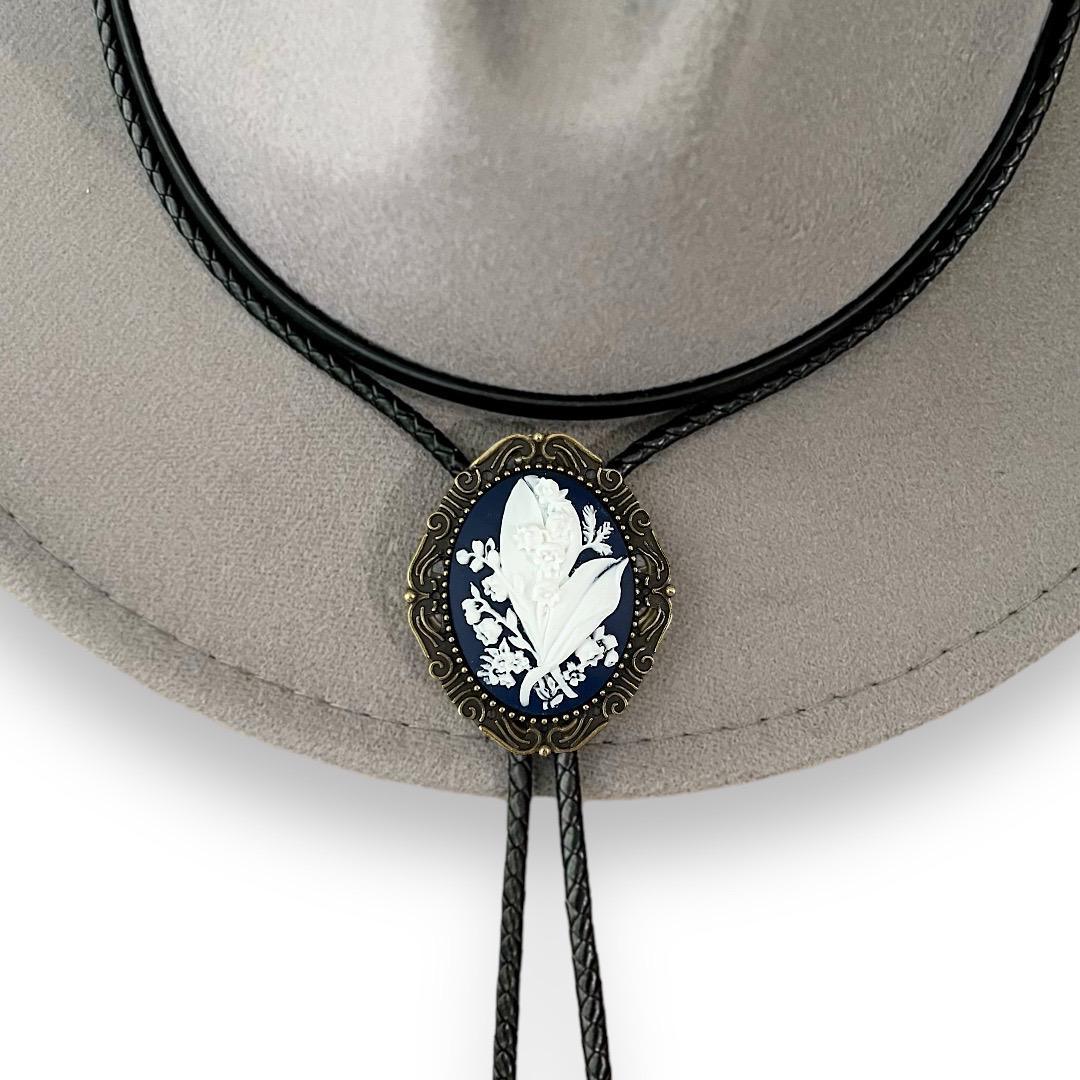 Infographic from The Bold Bow Tie titled Bolo Tie Features. Right: A vintage-style bolo tie with a blue and white cowboy riding a horse illustration inside an ornate bronze filigree frame on a black braided cord. Left: Lines connect the image to three checkmark features: Handmade, 39" Long Cord, and Pendant is 1.5" (L) x 1.25" (W). Bottom text reads Perfect Addition to Your Outfit.