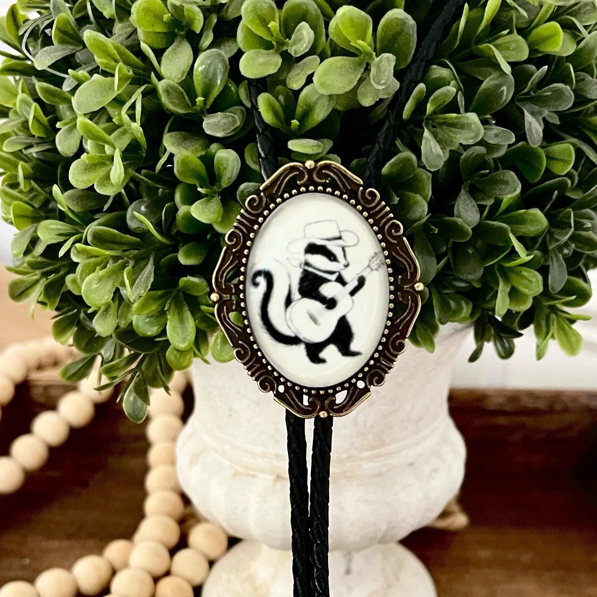 Infographic from The Bold Bow Tie titled Bolo Tie Features. Right: A vintage-style bolo tie with a blue and white cowboy riding a horse illustration inside an ornate bronze filigree frame on a black braided cord. Left: Lines connect the image to three checkmark features: Handmade, 39" Long Cord, and Pendant is 1.5" (L) x 1.25" (W). Bottom text reads Perfect Addition to Your Outfit.