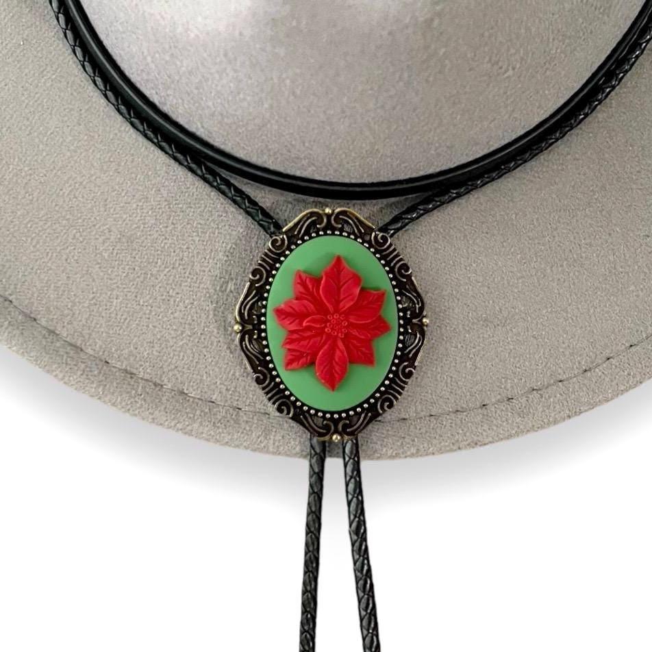 Infographic from The Bold Bow Tie titled Bolo Tie Features. Right: A vintage-style bolo tie with a blue and white cowboy riding a horse illustration inside an ornate bronze filigree frame on a black braided cord. Left: Lines connect the image to three checkmark features: Handmade, 39" Long Cord, and Pendant is 1.5" (L) x 1.25" (W). Bottom text reads Perfect Addition to Your Outfit.