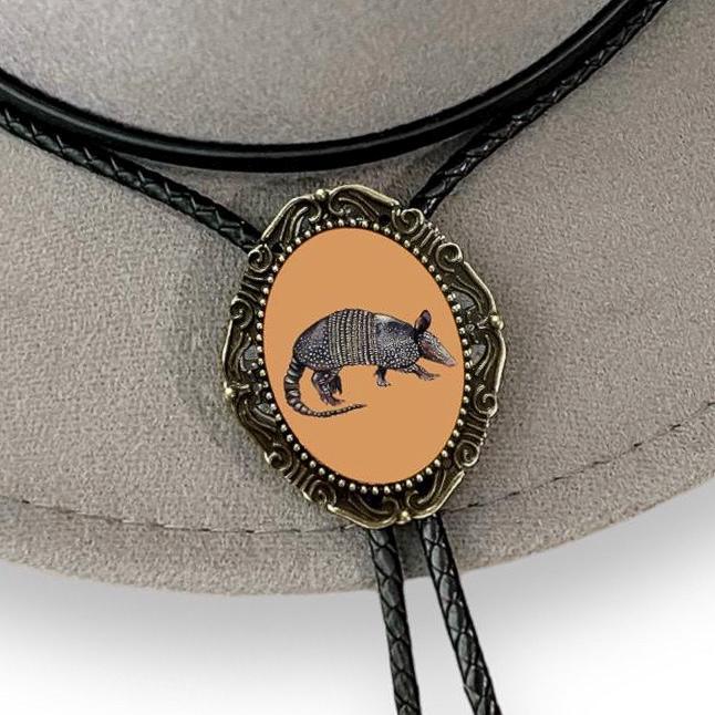 Close-up of a decorative bolo tie with an orange oval pendant featuring an armadillo, set on a beige hat.