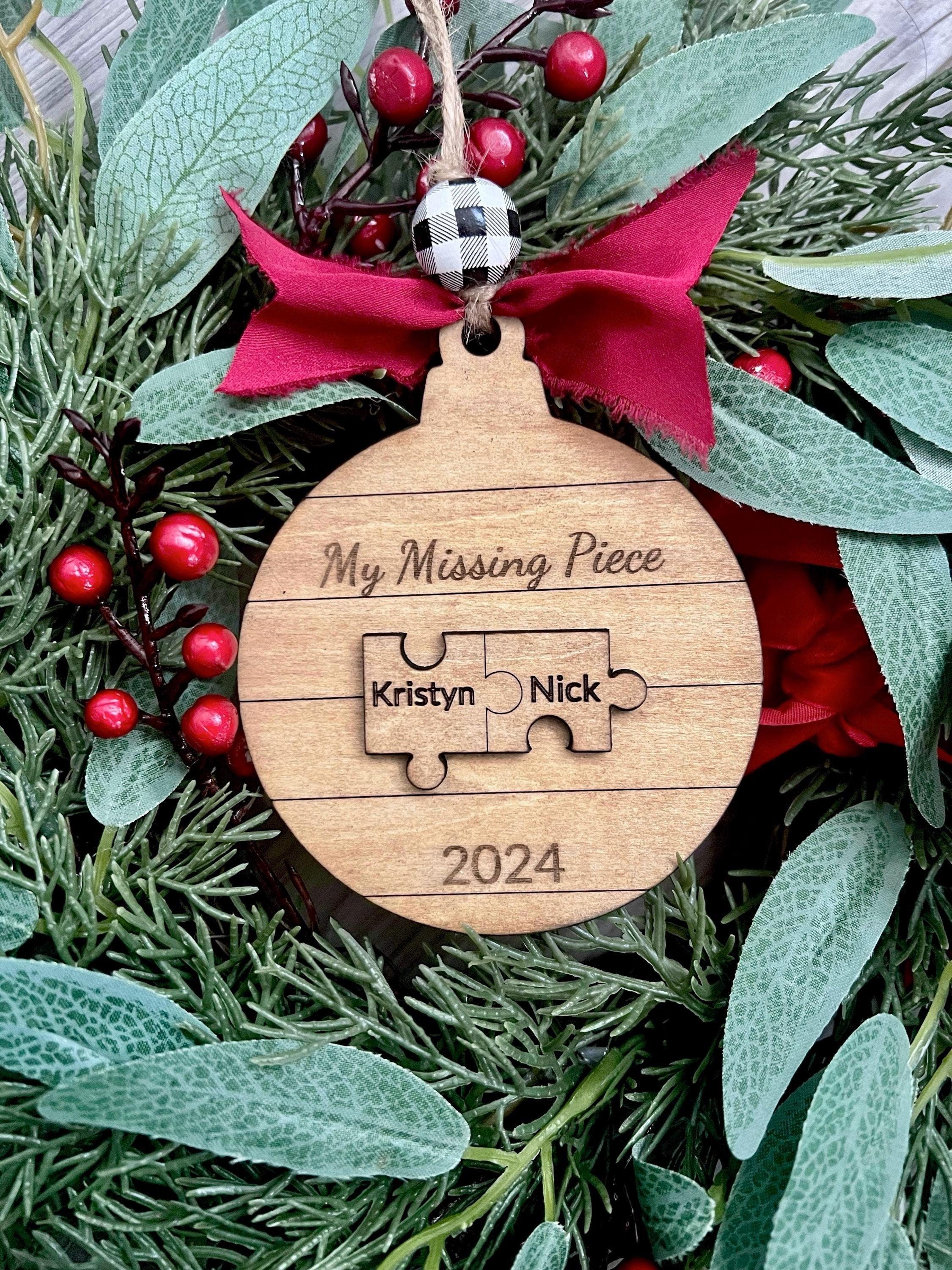 A wooden Christmas ornament with the text 'MY MISSING PIECE' and two puzzle pieces with names. It is adorned with a red bow, green leaves, and red berries.
