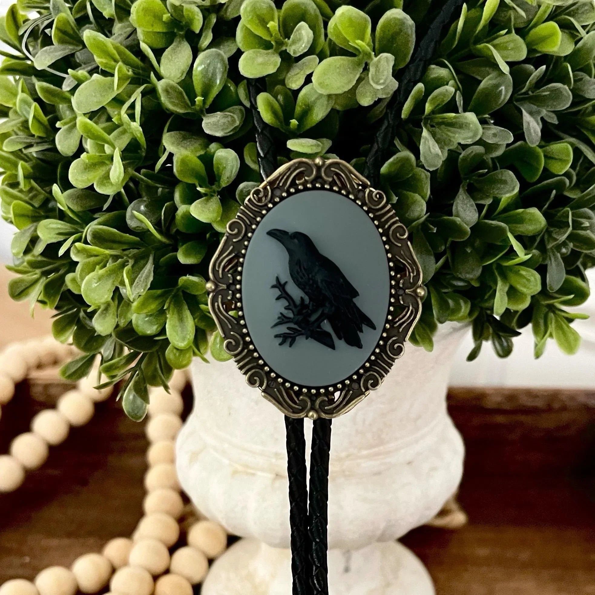 Crow Bolo tie draped among green foliage with the gray oval centerpiece and crow silhouette visible, braided leather cords.